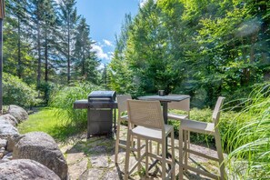 An outdoor patio with a grill, table, and chairs, surrounded by a forested area. - Tremblant Prestige - Les Falaises 138-2 (Mont-Tremblant)