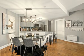 A modern kitchen with a central island, stainless steel appliances, and a dining area with a glass table and chairs. - Tremblant Prestige - Les Falaises 138-2 (Mont-Tremblant)