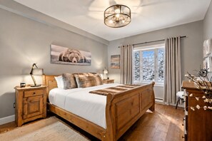 A bedroom with a wooden bed, a nightstand, a lamp, a window with curtains, and a wall-mounted artwork. - Tremblant Prestige - Les Falaises 138-2 (Mont-Tremblant)