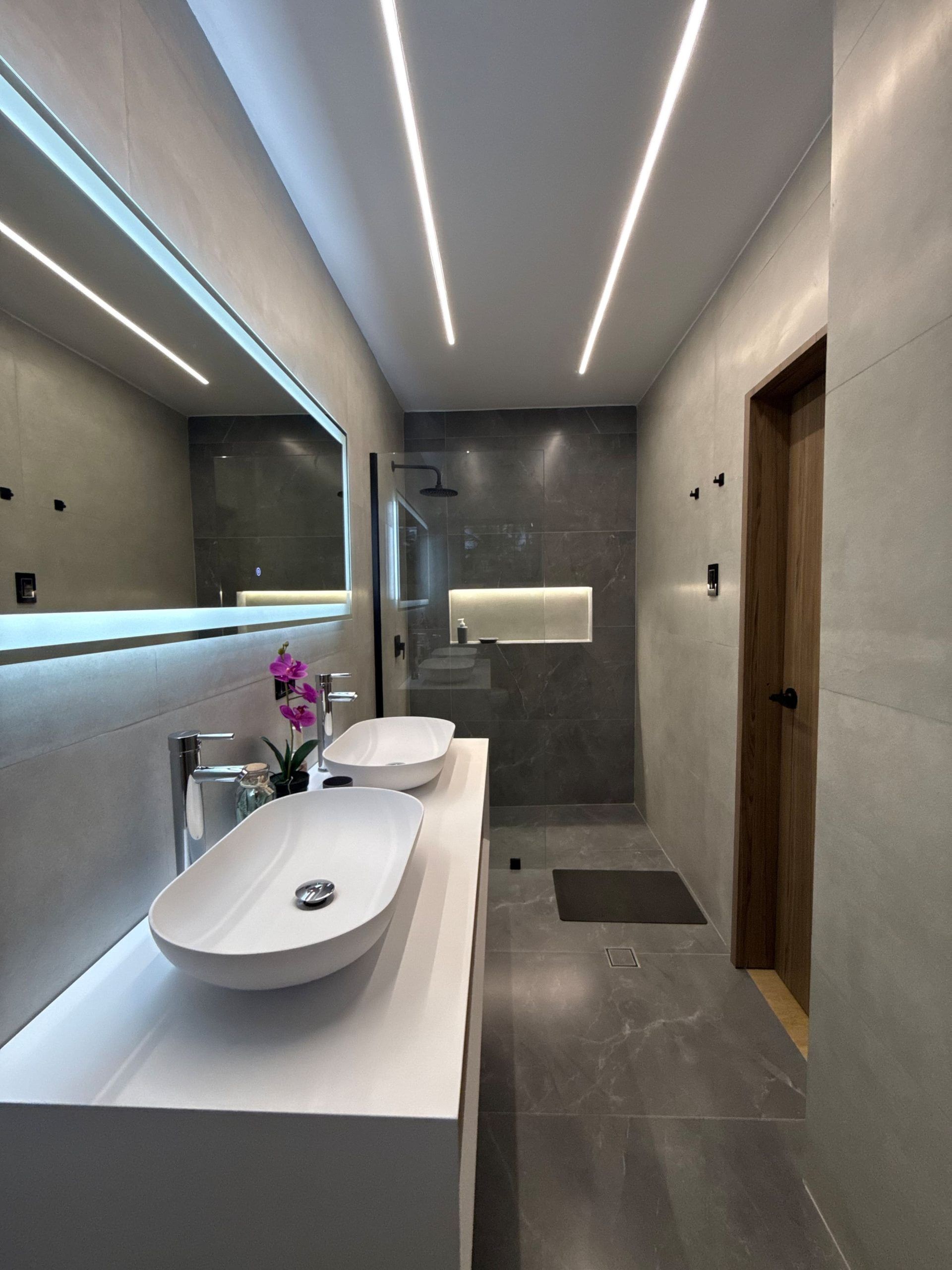 Een moderne badkamer met een grote spiegel, twee wastafels, een douche en een houten deur.