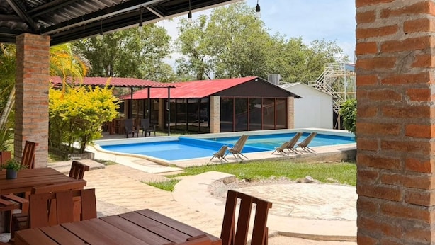An outdoor pool area with a covered patio, wooden furniture, and a building with a red roof in the background. - Hotel y Centro Turístico Valle Encantado (Gracias)