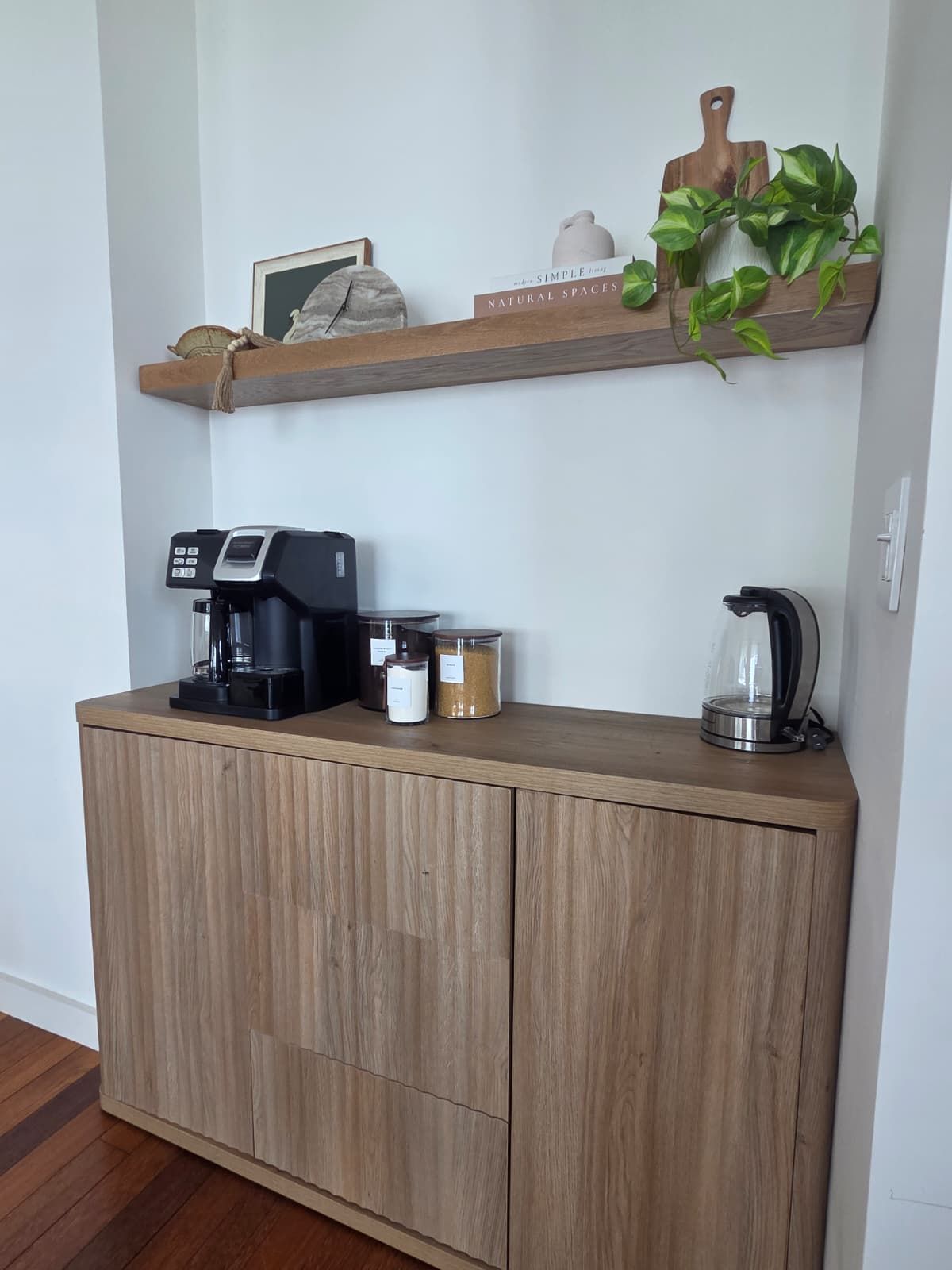 Une petite cuisine équipée d’une machine à café, de bocaux et d’une bouilloire, le tout posé sur un meuble en bois.