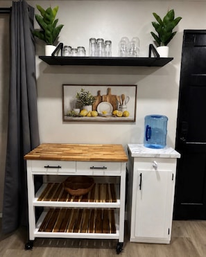 Eine Küchenzeile mit einem weißen Schrank, einem Holztisch mit Schubladen, einem Regal für Gläser, einem Wasserspender und einem gerahmten Bild mit Küchenutensilien.