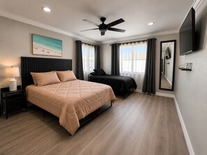 Quarto com cama, mesas de cabeceira, ventoinha no teto, televisão e janela com cortinas.