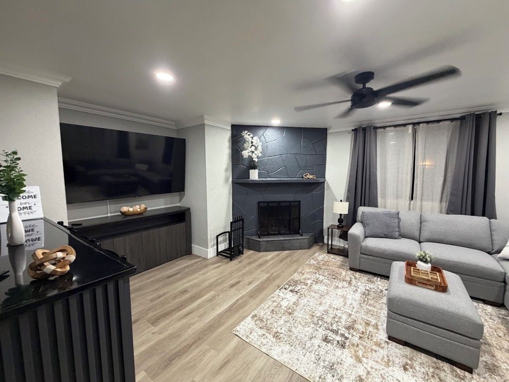 A modern living room with a flat-screen TV, fireplace, grey sofa, ottoman, and a ceiling fan.