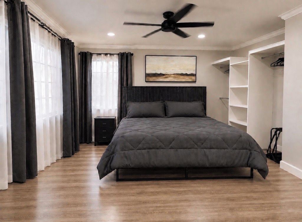 A bedroom with a bed, a ceiling fan, a nightstand, a wardrobe, and a window with curtains.