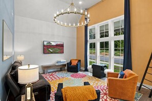 Une chambre d’hôtel dotée d’une grande fenêtre, d’un lustre, d’un canapé, d’un fauteuil et d’un téléviseur fixé au mur.