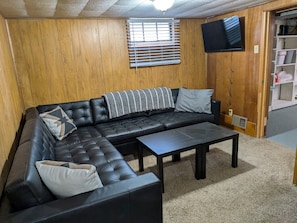 A room with a sectional sofa, a coffee table, wooden walls, a TV mounted on the wall, and a window with blinds.