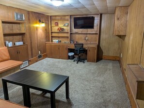 Ein Zimmer mit Schreibtisch aus Holz, einem an der Wand befestigten Fernseher, einer Sitzecke mit Polstern und einer kleinen Mülleimer.