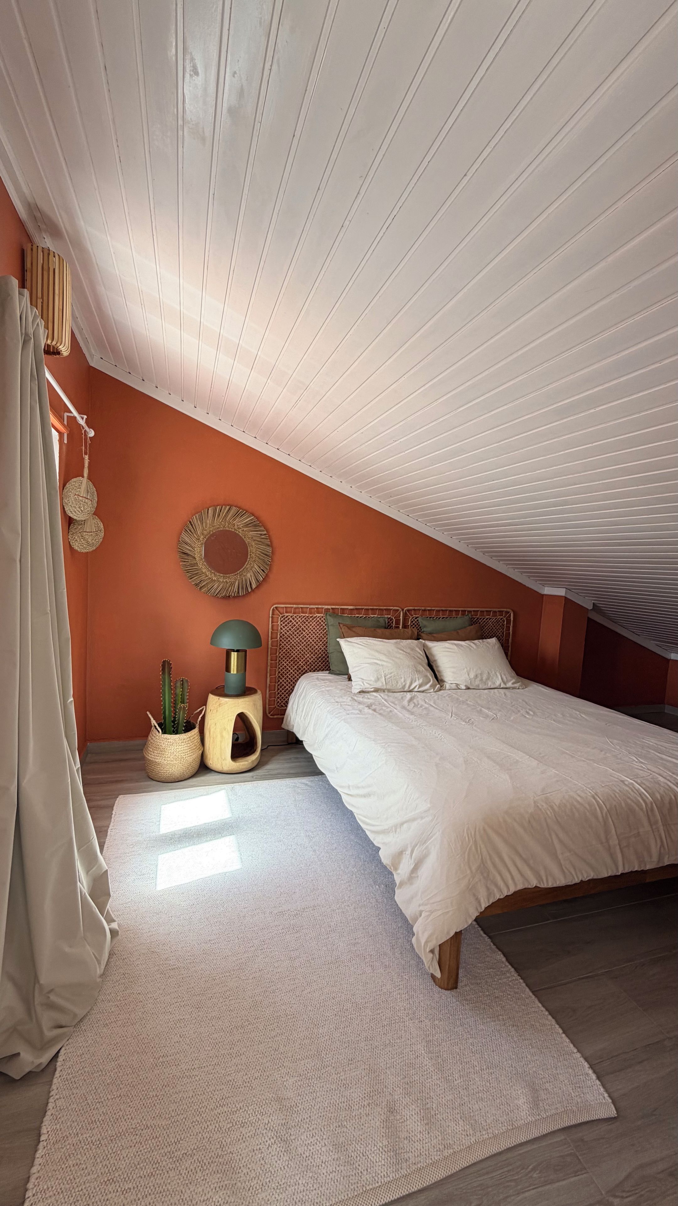 Ein Schlafzimmer mit schräg steilender Decke, einem Bett mit weißer Bettwäsche, einem Holzkopfende, einem kleinen Nachttisch mit Lampe, einem an der Wand befestigten Spiegel und einer Kakteenpflanze.
