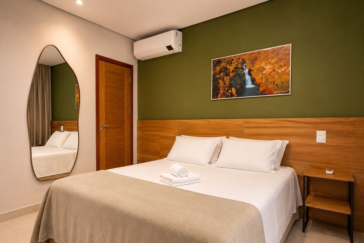 A hotel room with a bed, a wooden headboard, a wall-mounted air conditioner, a painting, a mirror, and a bedside table.