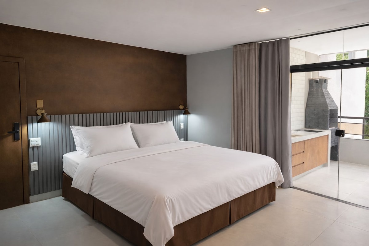 A modern bedroom with a large bed, a wooden headboard, a built-in bench, and a sliding glass door leading to a balcony with a stone fireplace.