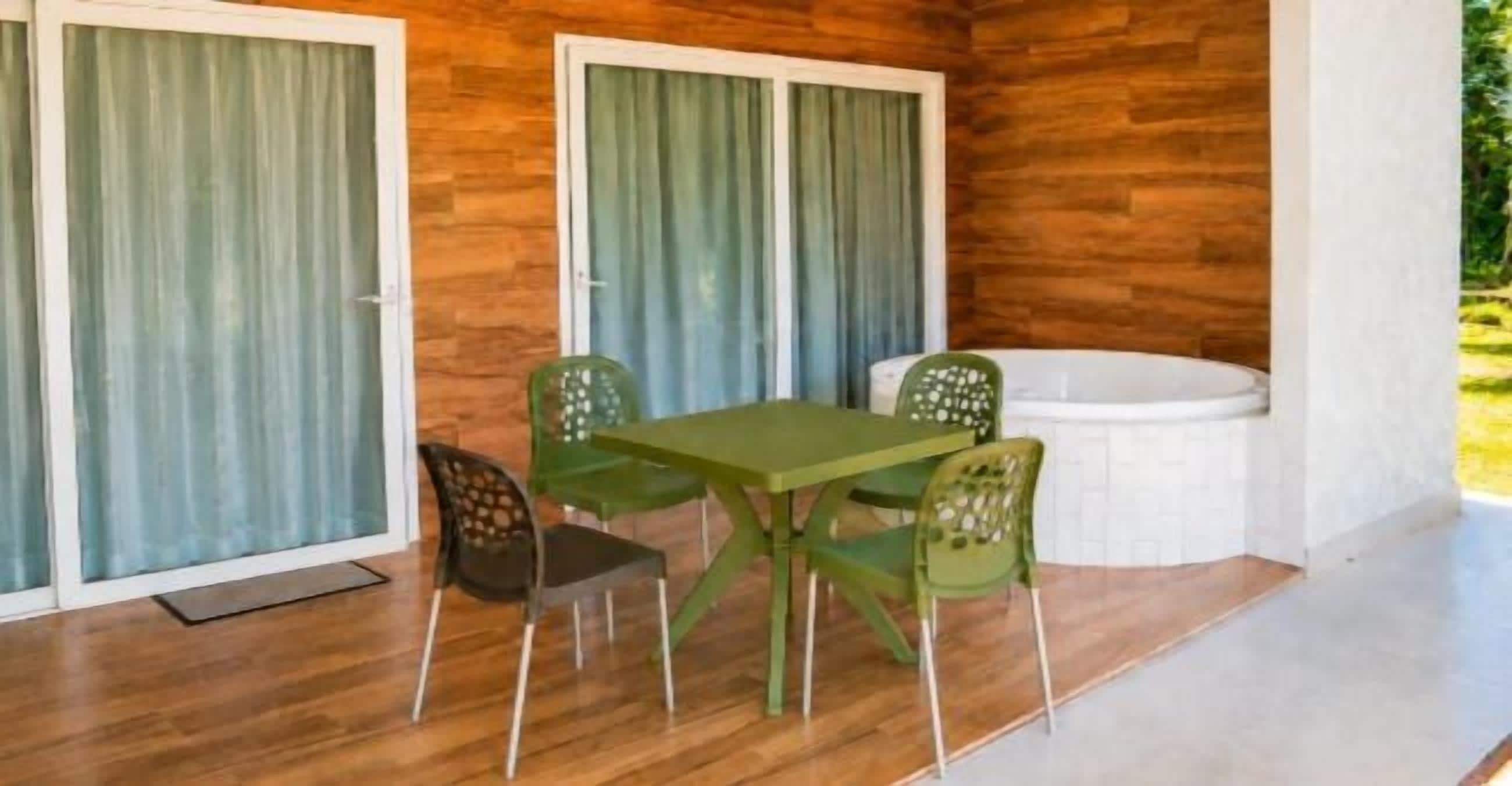 A room with a wooden floor, a green table, four chairs, a white bathtub, and sliding glass doors with curtains.