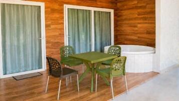 A room with a wooden floor, a green table, four chairs, a white bathtub, and sliding glass doors with curtains.