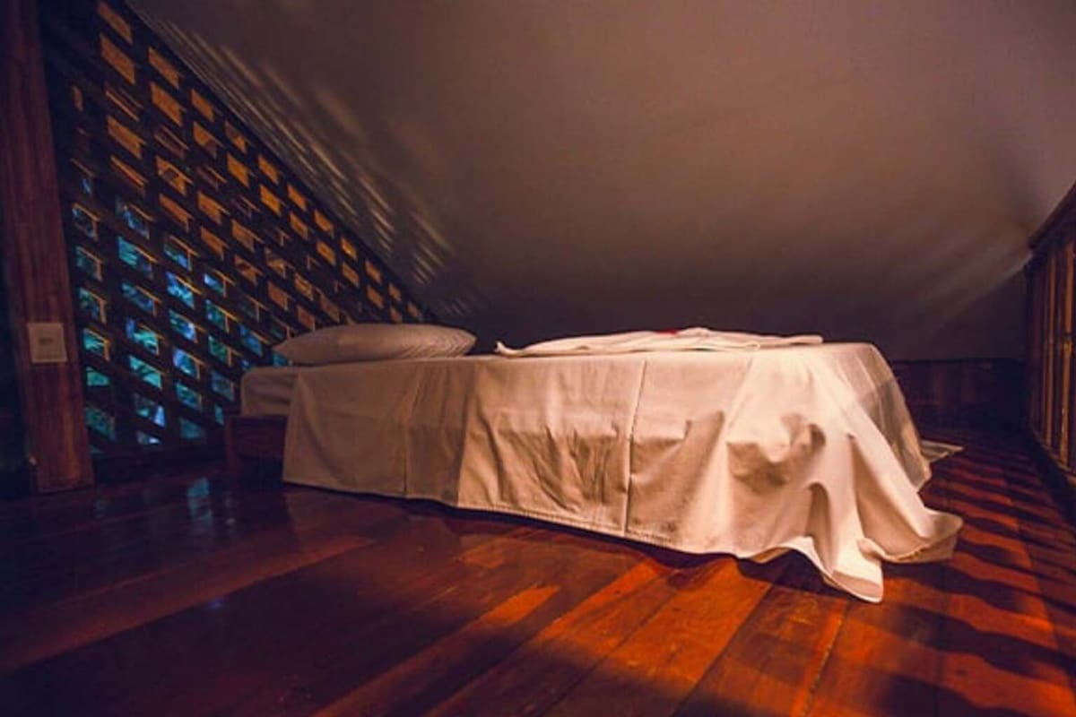 A room with a single bed, a wooden floor, and a wall of illuminated windows.