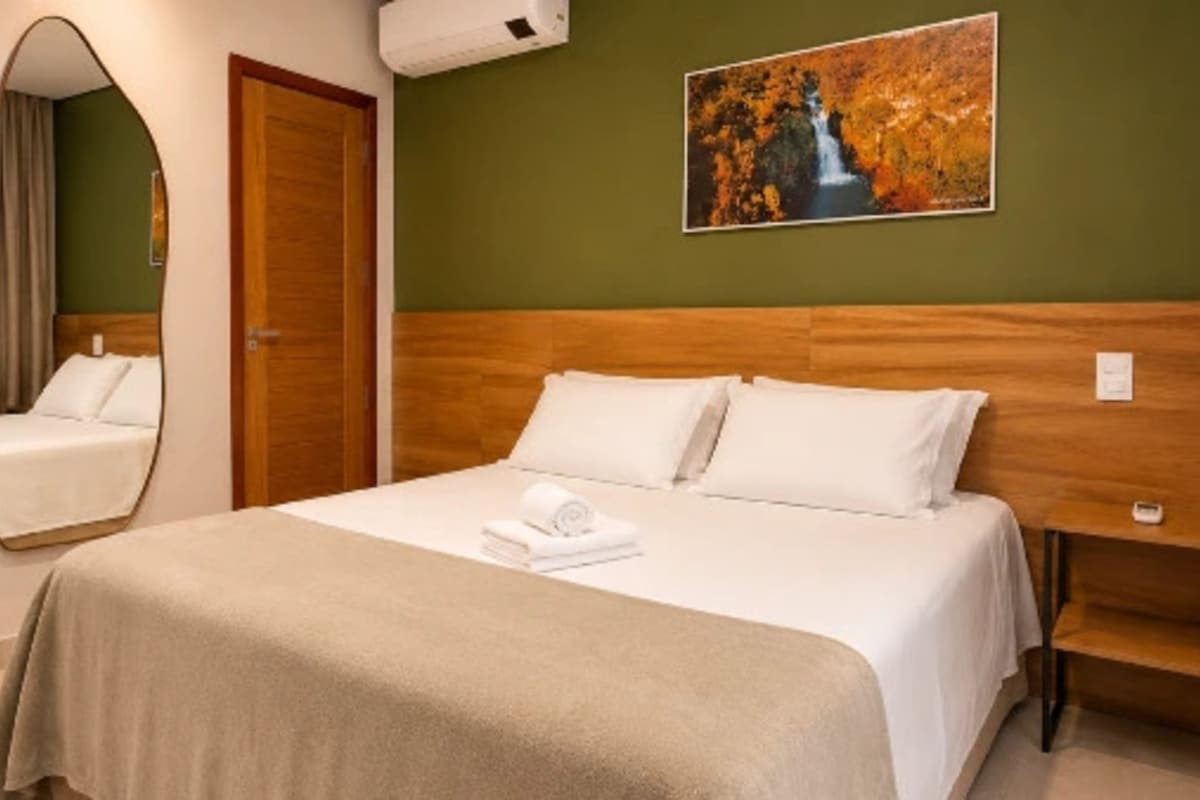A hotel room with a bed, a wooden headboard, a painting, a mirror, and a bench.