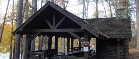 Una cabaña de madera con una mesa de picnic cubierta y bancos, rodeada de árboles.