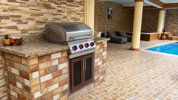 An outdoor kitchen with a built-in grill and oven, a pool in the background, and a stone-tiled floor. - Casa Nube | 8 min to SDQ Airport | Private Pool & Jacuzzi (Santo Domingo Este)