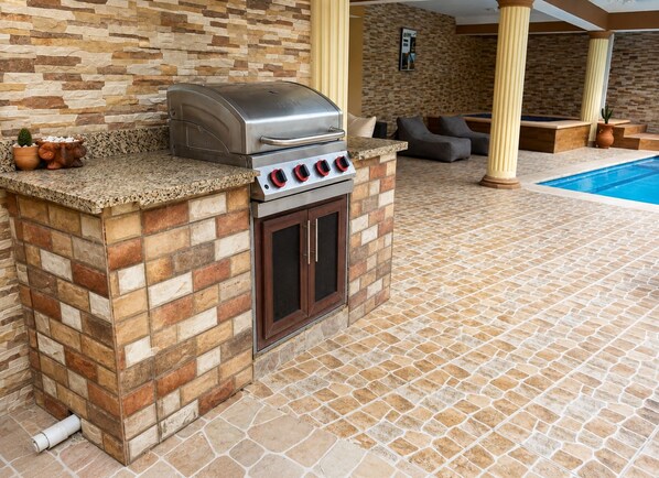 Eine Außenküche mit integriertem Grill und Backofen, im Hintergrund ein Pool und ein Boden mit Steinplatten.