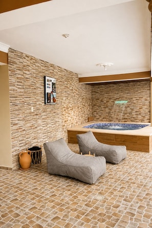 A spa area with a hot tub, two grey lounge chairs, a tiled floor, and a wall with a textured pattern. - Casa Nube | 8 min to SDQ Airport | Private Pool & Jacuzzi (Santo Domingo Este)