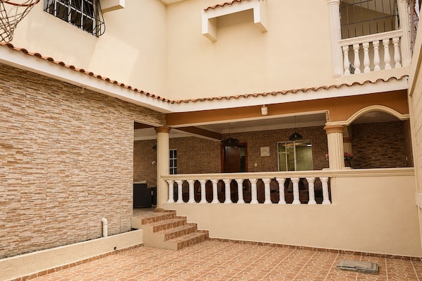 A covered outdoor area with a brick wall, a balcony with a balustrade, and a tiled floor.