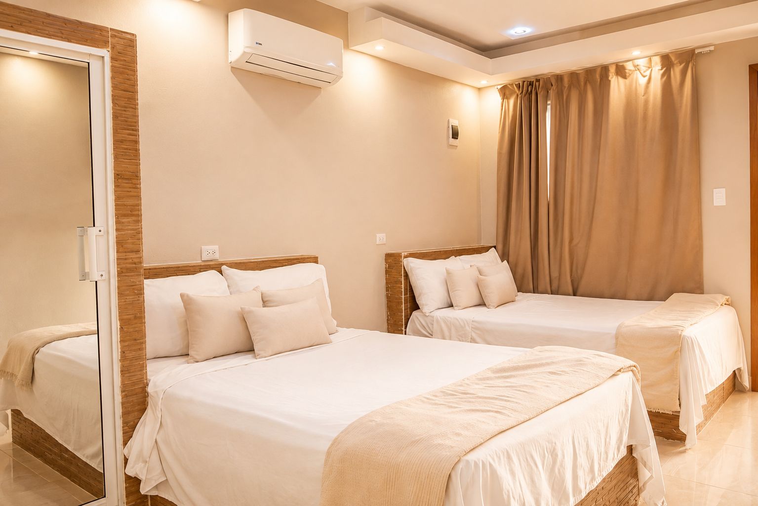 A hotel room with two beds, a wooden headboard, beige bedding, an air conditioning unit, and a window with curtains.