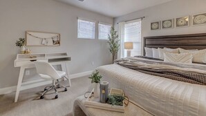 Ein Schlafzimmer mit Bett, Schreibtisch und Stuhl. Es gibt Fenster mit Jalousien, eine Wandregal mit Dekoration und einen Nachttisch mit Lampe und Pflanze.