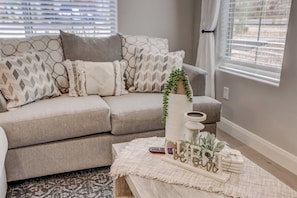 A living room with a grey sofa, patterned cushions, a coffee table with decorative items, and a window with blinds. - Hot Tub, Heated Pool with Slide, Pickleball, Bunk Beds,  2PM Check Out (Washington)