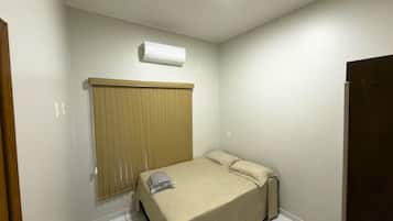 Una habitación pequeña con una cama individual, cabecera de madera, unidad de aire acondicionado blanca en la pared y una puerta cerrada.