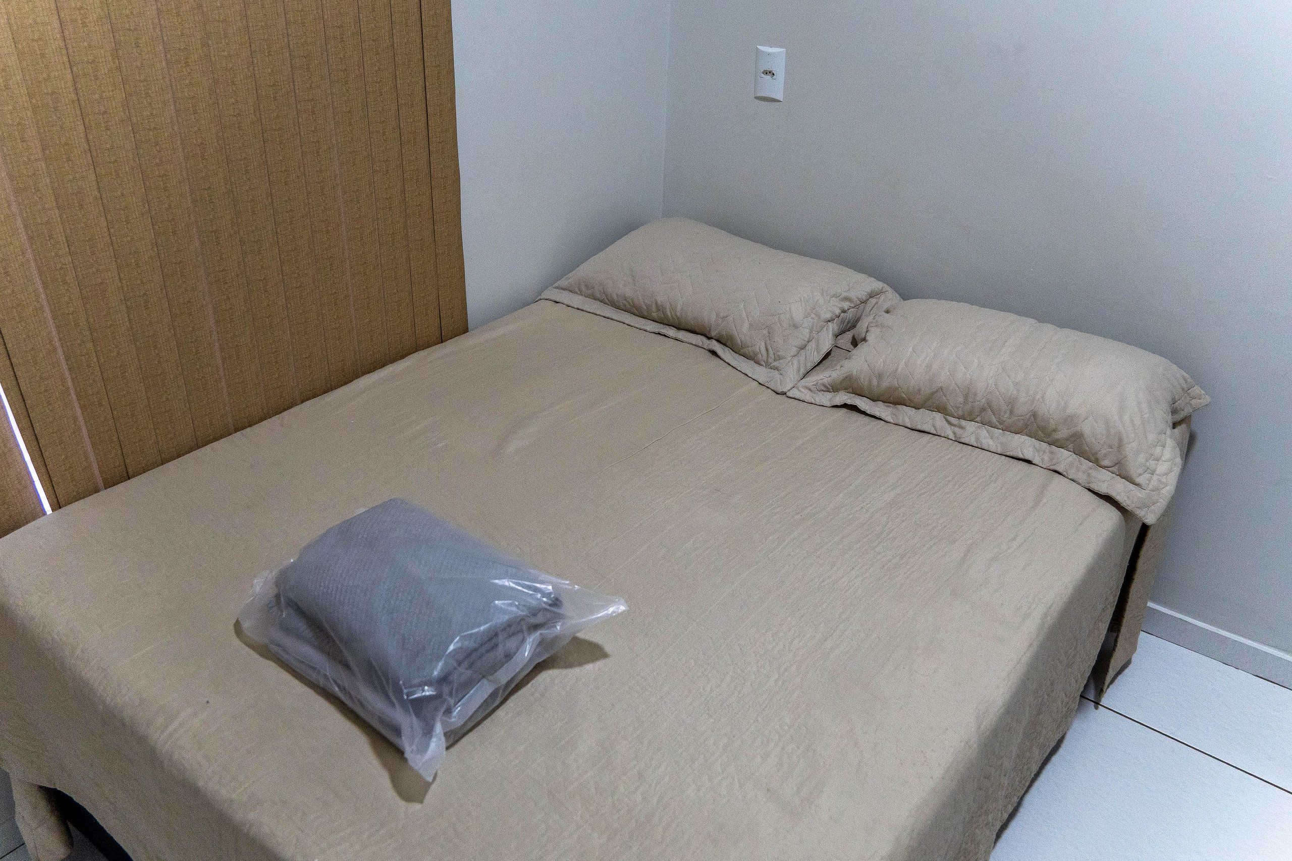 Una habitación pequeña con una cama individual, colchón y almohadas color beige, una bolsa de plástico sobre la cama y una puerta de madera a la izquierda.