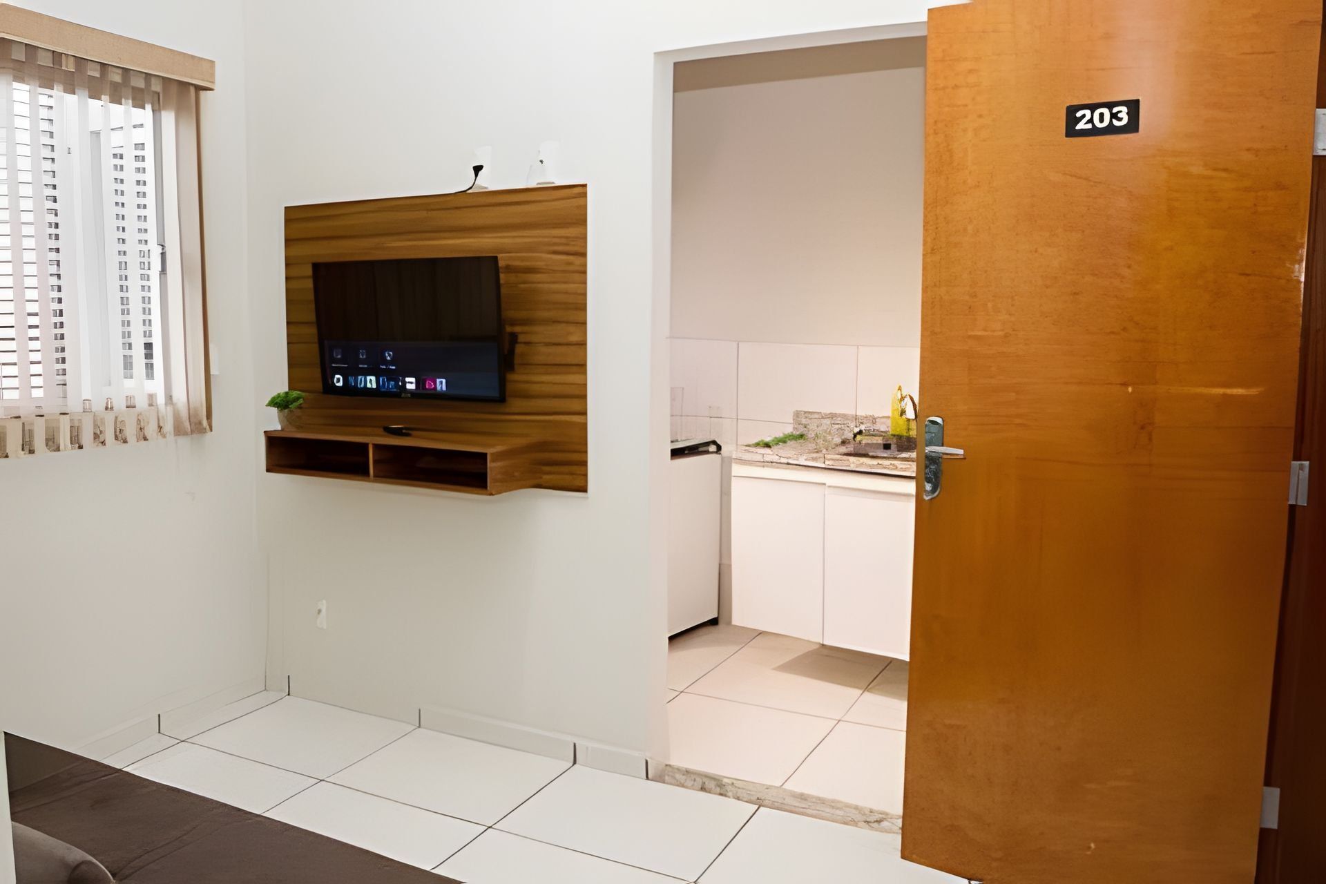 Una habitación con televisión montada, una puerta con la etiqueta '203' y una vista parcial de la cocina.