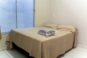A single bed with a beige bedspread and a plastic bag on it, in a room with a tiled floor and a closed door.
