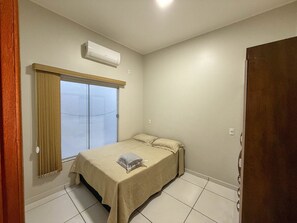 A bedroom with a bed, a window with curtains, an air conditioning unit, and a door. - CONDOMÍNIO SONHARE - 103 (ANAPOLIS)
