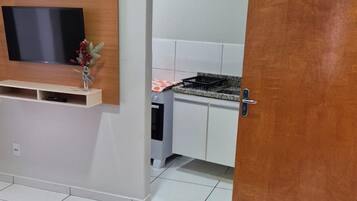 Una habitación con sofá, televisión instalada en un panel de madera, zona de cocina con estufa y armarios, y una puerta con el número 103.