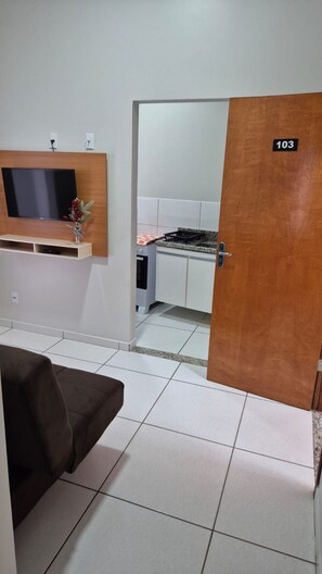 A room with a sofa, a television mounted on a wooden panel, a kitchen area with a stove and cabinets, and a door with the number 103. - CONDOMÍNIO SONHARE - 103 (ANAPOLIS)
