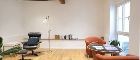 A room with a wooden floor, a black leather chair, an orange chair, a small table with books, and a floor lamp.