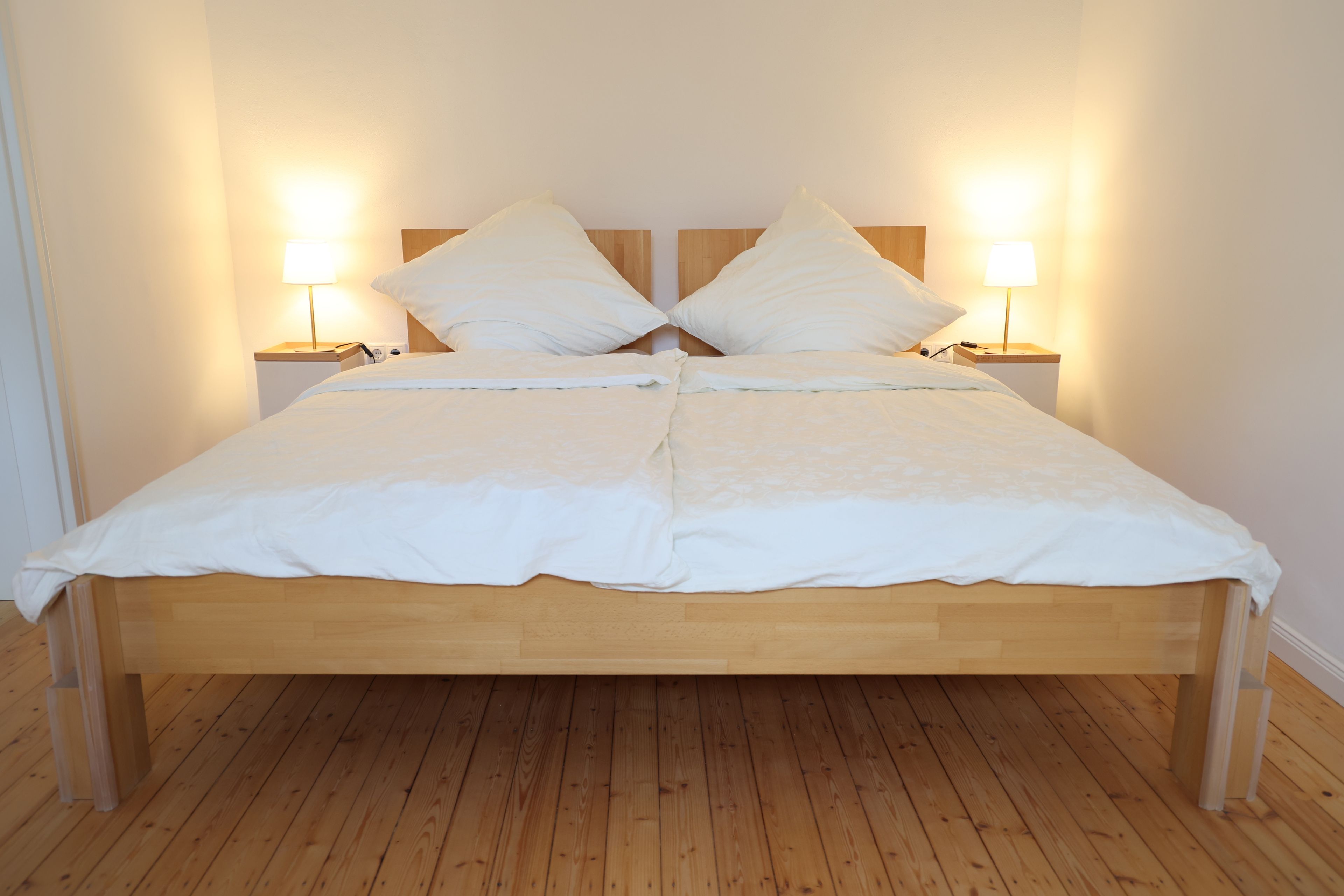 Ein Doppelbett mit Holzrahmen und weißer Bettwäsche, flankiert von zwei Nachttischen mit Lampen, in einem Raum mit Holzboden.