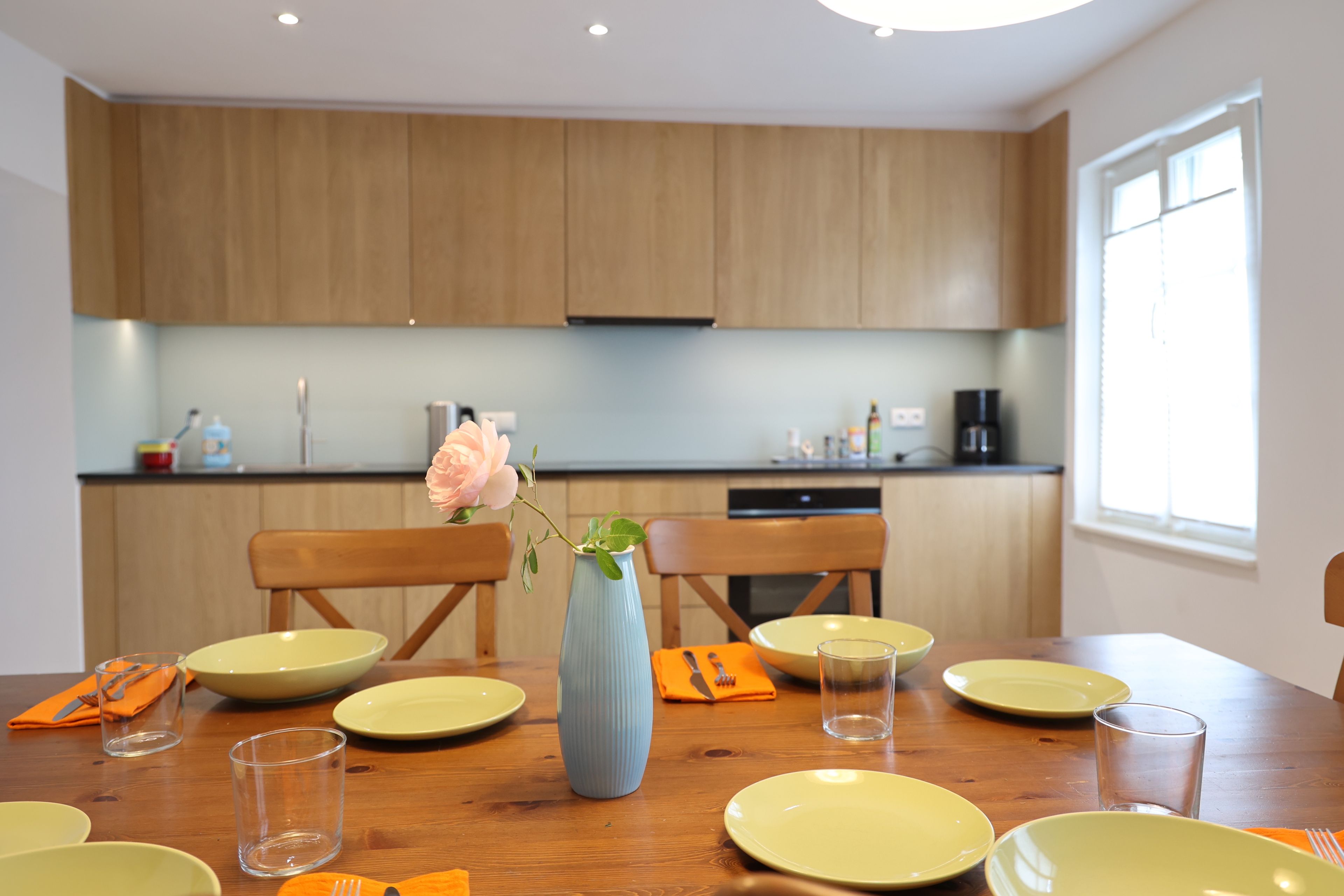 Een moderne keuken met houten kastjes, een eettafel gedekt met gele borden, en een vaas met een bloem.