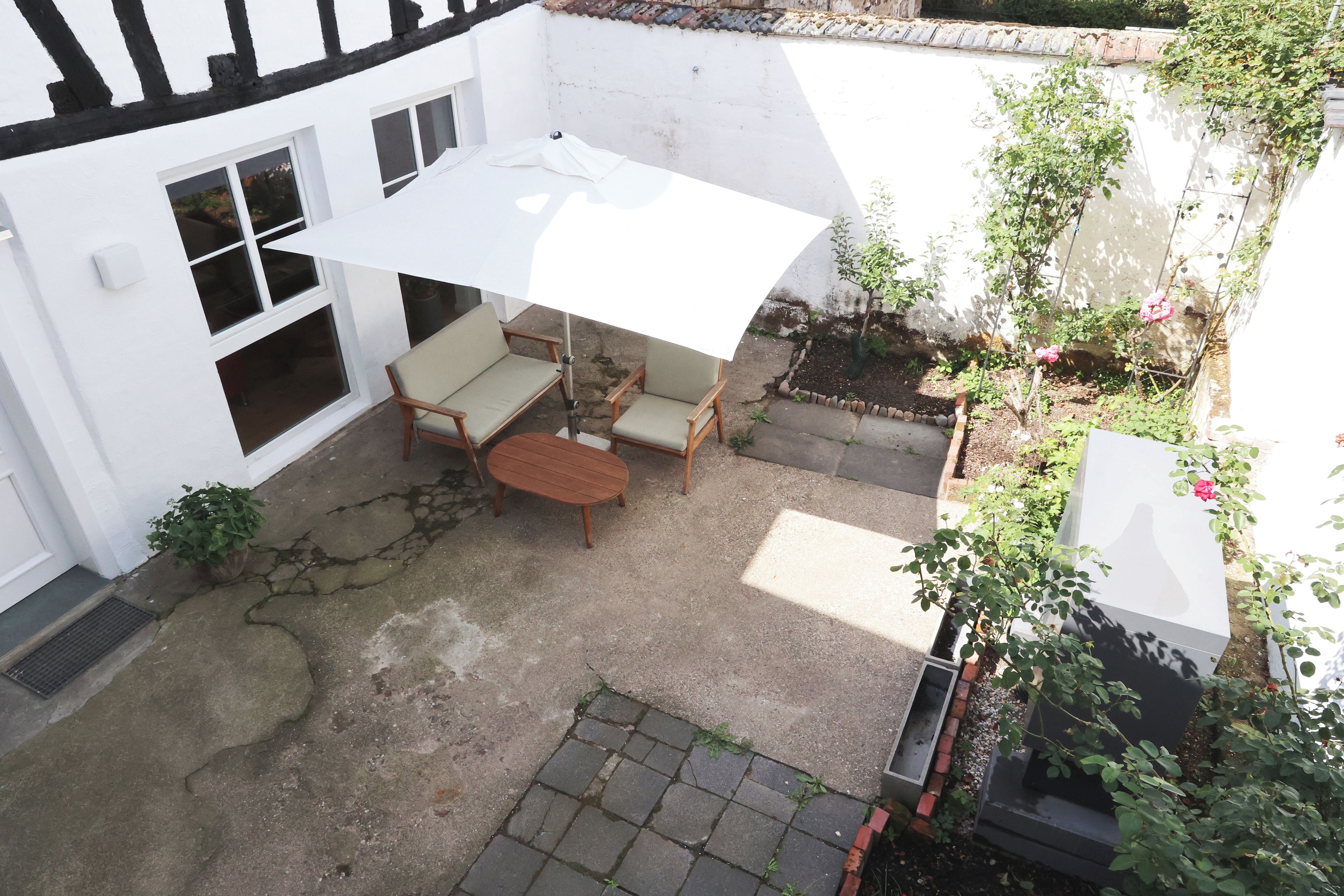 Een terras met een tafel en stoelen, een witte parasol, en een tuin met planten en een bakstenen rand.