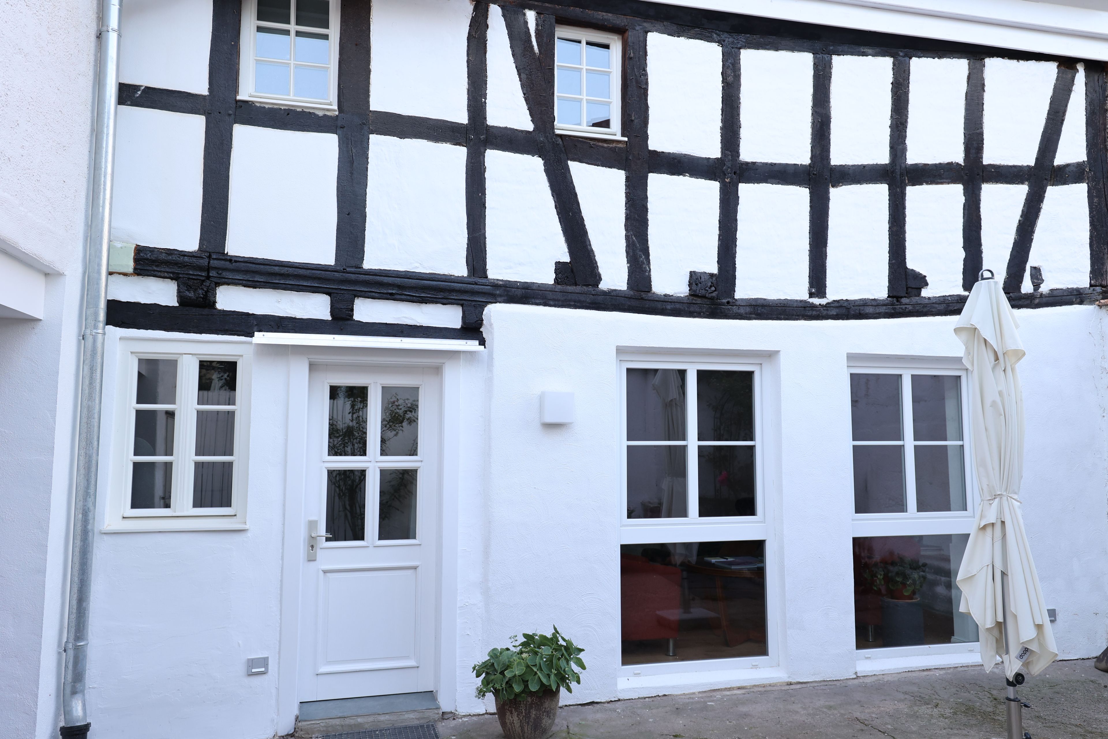A traditional half-timbered house with white walls and black timber framing. There are two windows with white frames, one door, and a small potted plant in front.