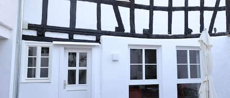 A traditional half-timbered house with white walls and black timber framing. There are two windows with white frames, one door, and a small potted plant in front.