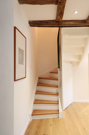 Eine Treppe mit hölzernen Stufen führt zu einer Zwischenebene, an der Wand hängt ein gerahmtes Bild und die Decke zeigt freiliegende Holzbalken.