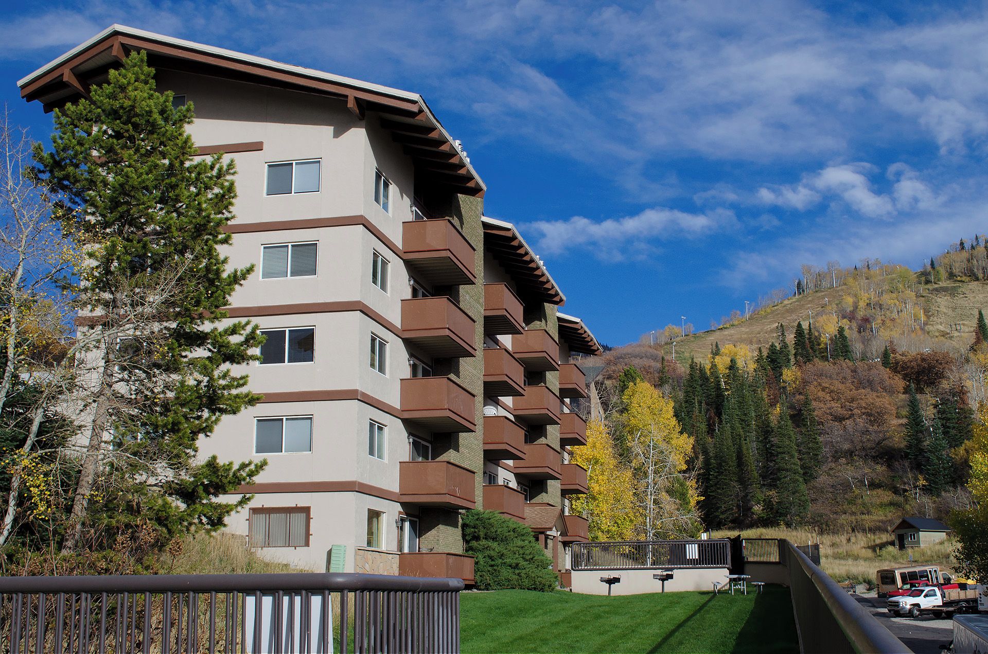 A multi-story residential building with balconies, surrounded by trees and a hilly landscape.