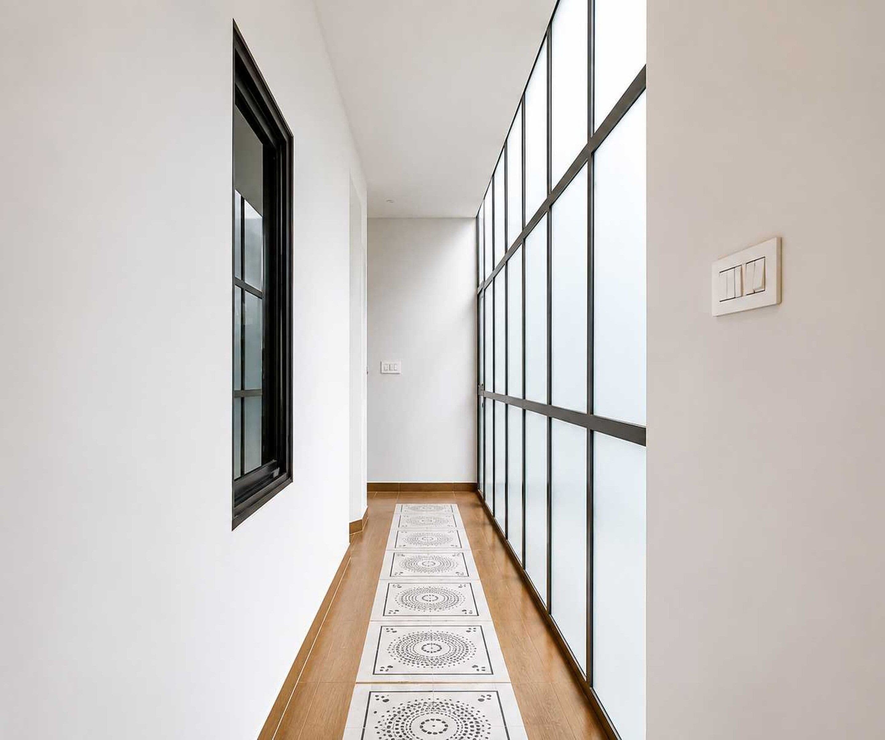 A corridor with a window on the left, a light switch on the right, and patterned floor tiles.