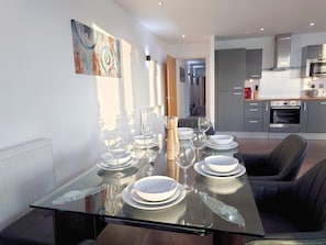 A modern dining area with a glass table set for six, a kitchen in the background, and a painting on the wall. - Modern 2-Bedroom Apartment Near ExCeL London (London)