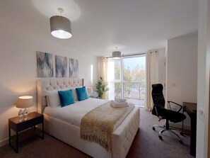 A bedroom with a large bed, a desk, and a chair. There is a window with curtains, a lamp on the bedside table, and a painting on the wall. - Modern 2-Bedroom Apartment Near ExCeL London (London)