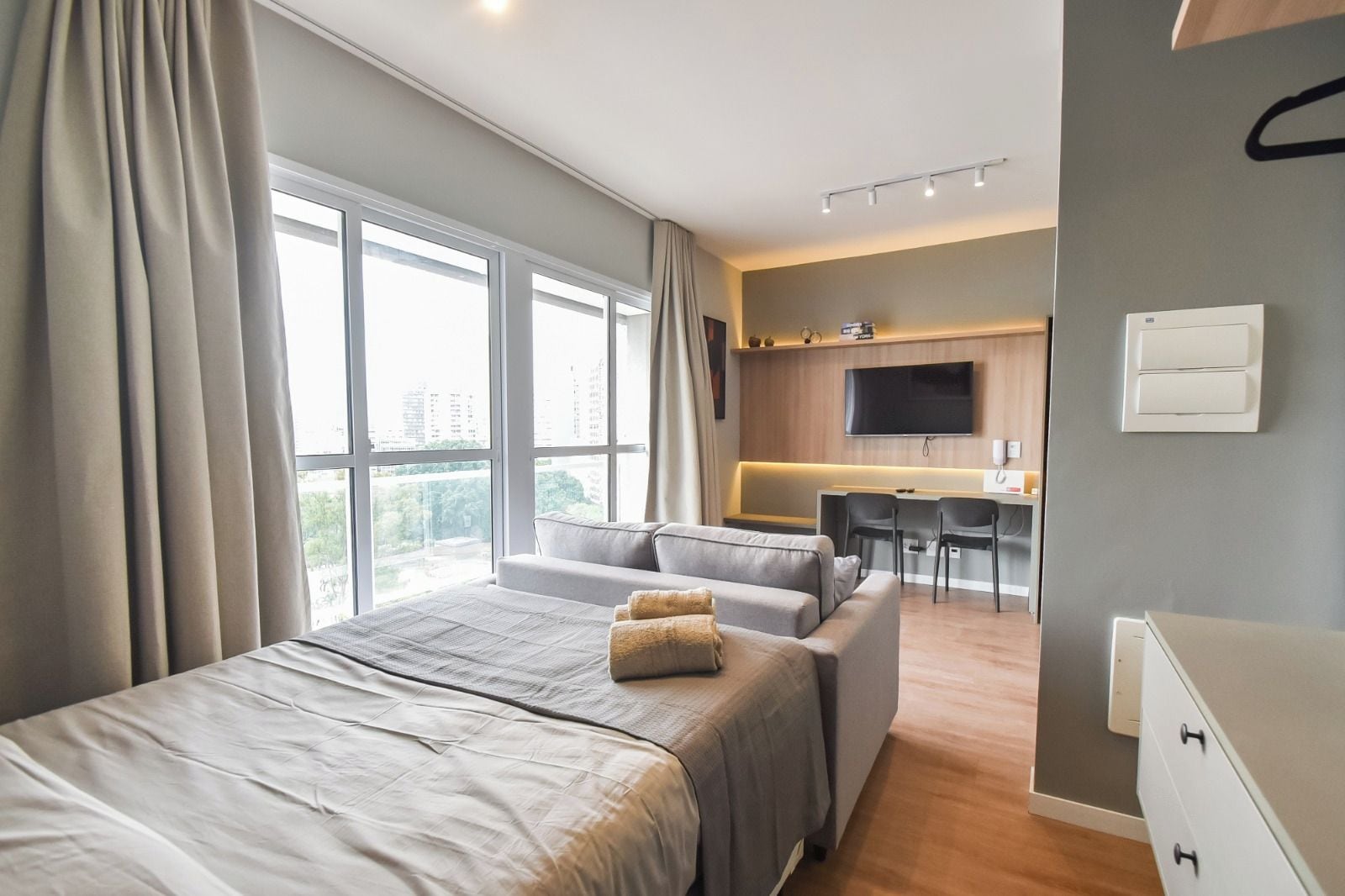 A modern hotel room with a large bed, a sofa, a desk with chairs, a flat-screen TV, and a city view.