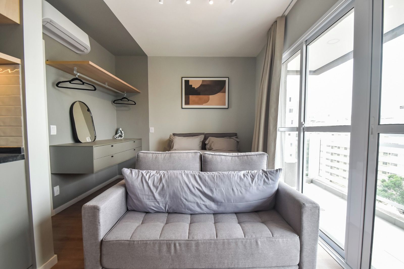 Um quarto moderno com um sofá cinzento, uma cama com almofadas, um espelho fixado na parede e um quadro.