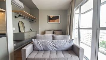Um quarto moderno com um sofá cinzento, uma cama com almofadas, um espelho fixado na parede e um quadro.