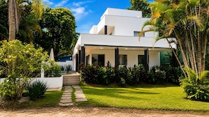 A modern two-story house with a paved driveway leading to a front door, surrounded by lush greenery and palm trees. - Villaggio 2 (São Sebastião)